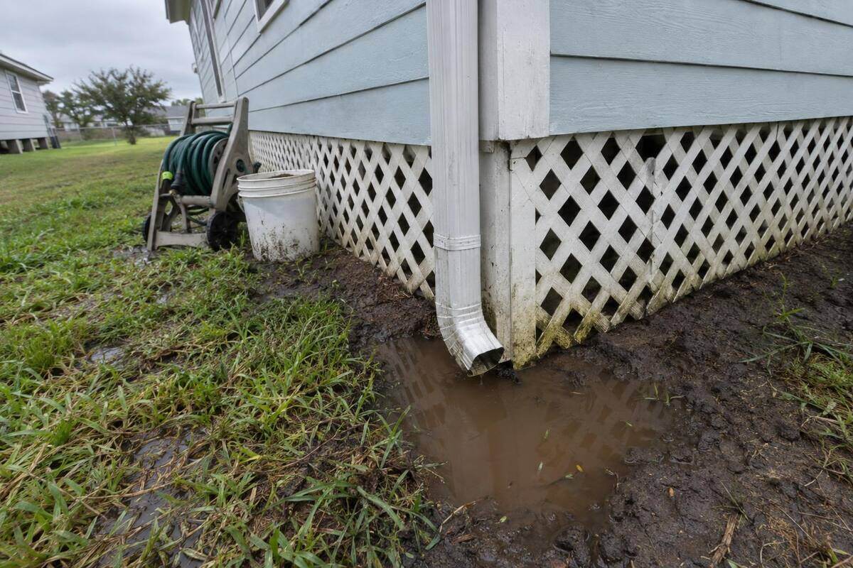 Aluminum downspout ending beside the lattice skirting of a pier-and-beam house with standing water on dark clay soil