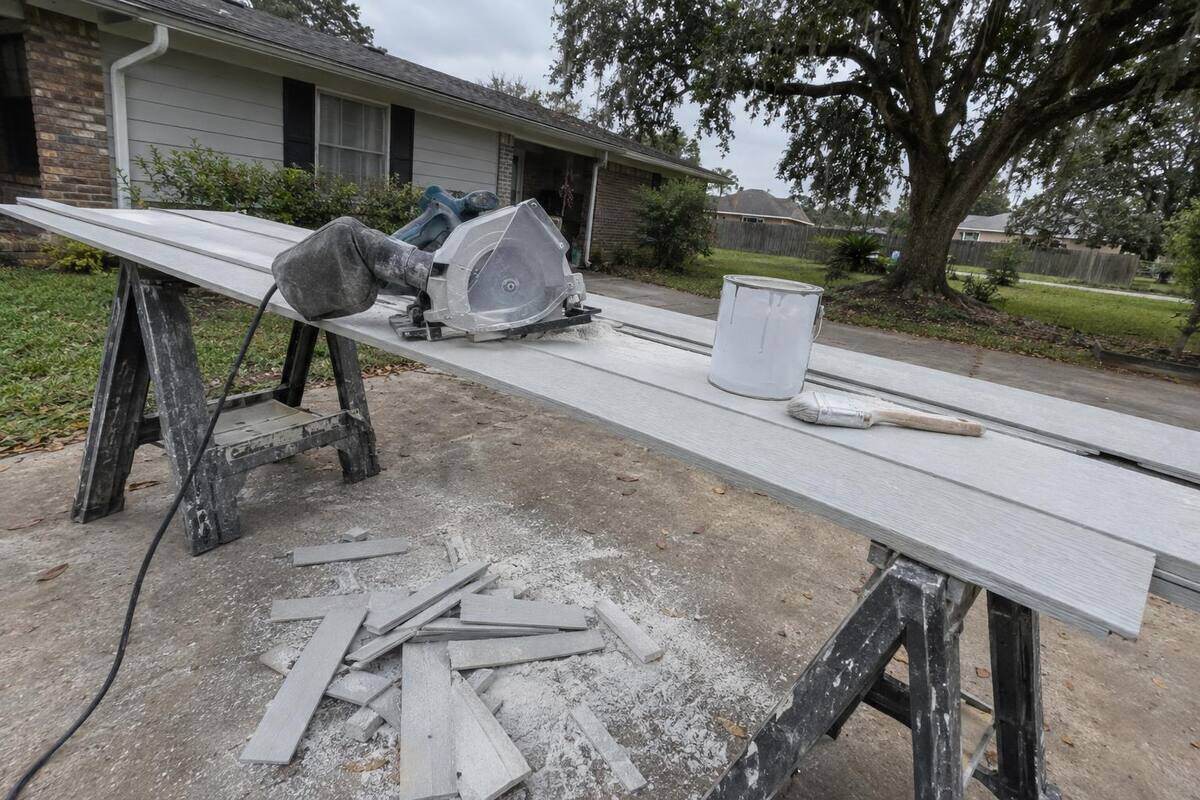 Fiber-cement lap planks laid across two sawhorses beside a dust-shrouded circular saw, with a stack of cut offcuts and a can of primer on the ground