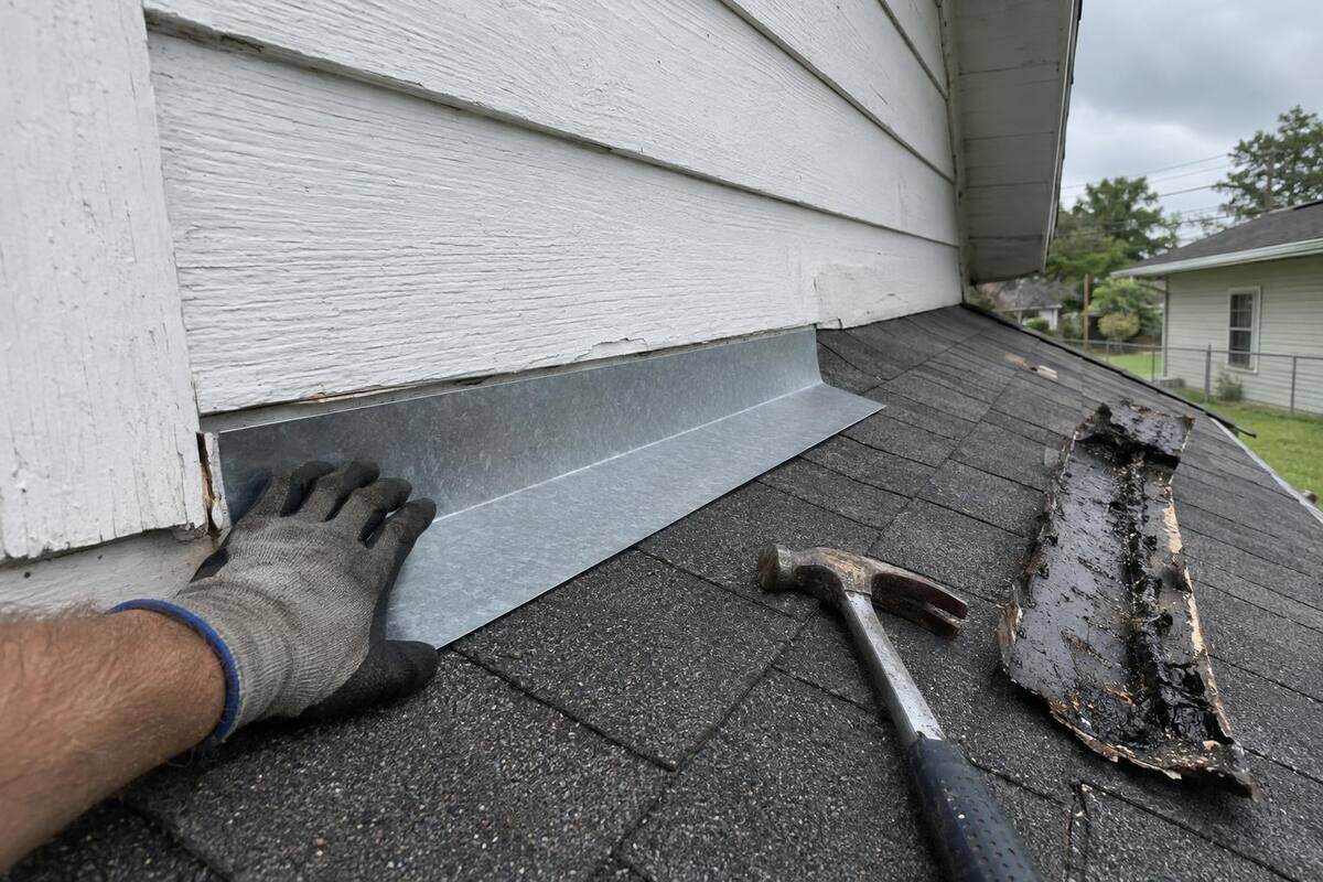 Step flashing being fitted where a low porch roof meets a painted wood-sided wall, with a hammer and a strip of old tar-covered flashing on the shingles
